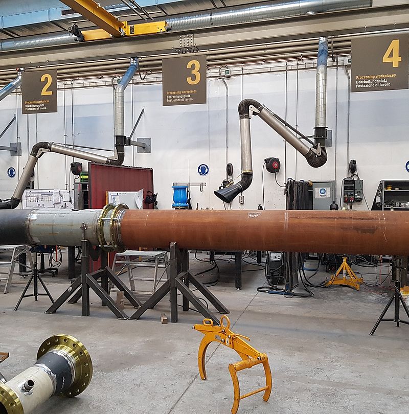 Workshop with long steel pipe on stands and numbered workstations with extraction systems.