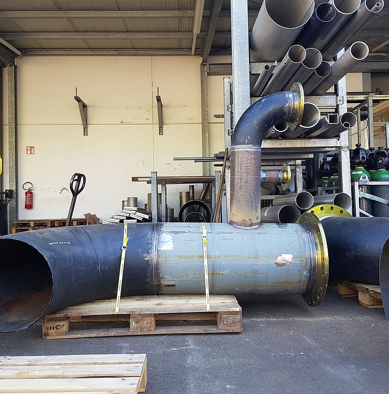 Large steel pipe with flanges and bend on pallets in workshop with pipe storage.