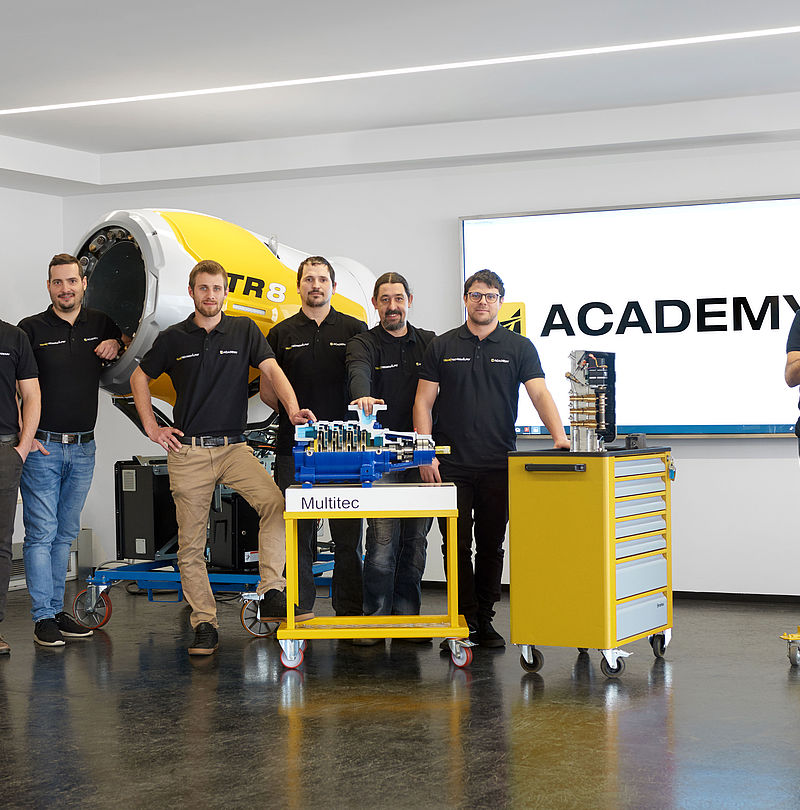 TechnoAlpin Academy team in black polo shirts in front of TR8 snow generator and training equipment.