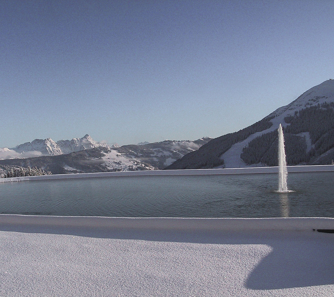 Speicherteich mit Fontäne und winterlichem Bergpanorama im Skigebiet