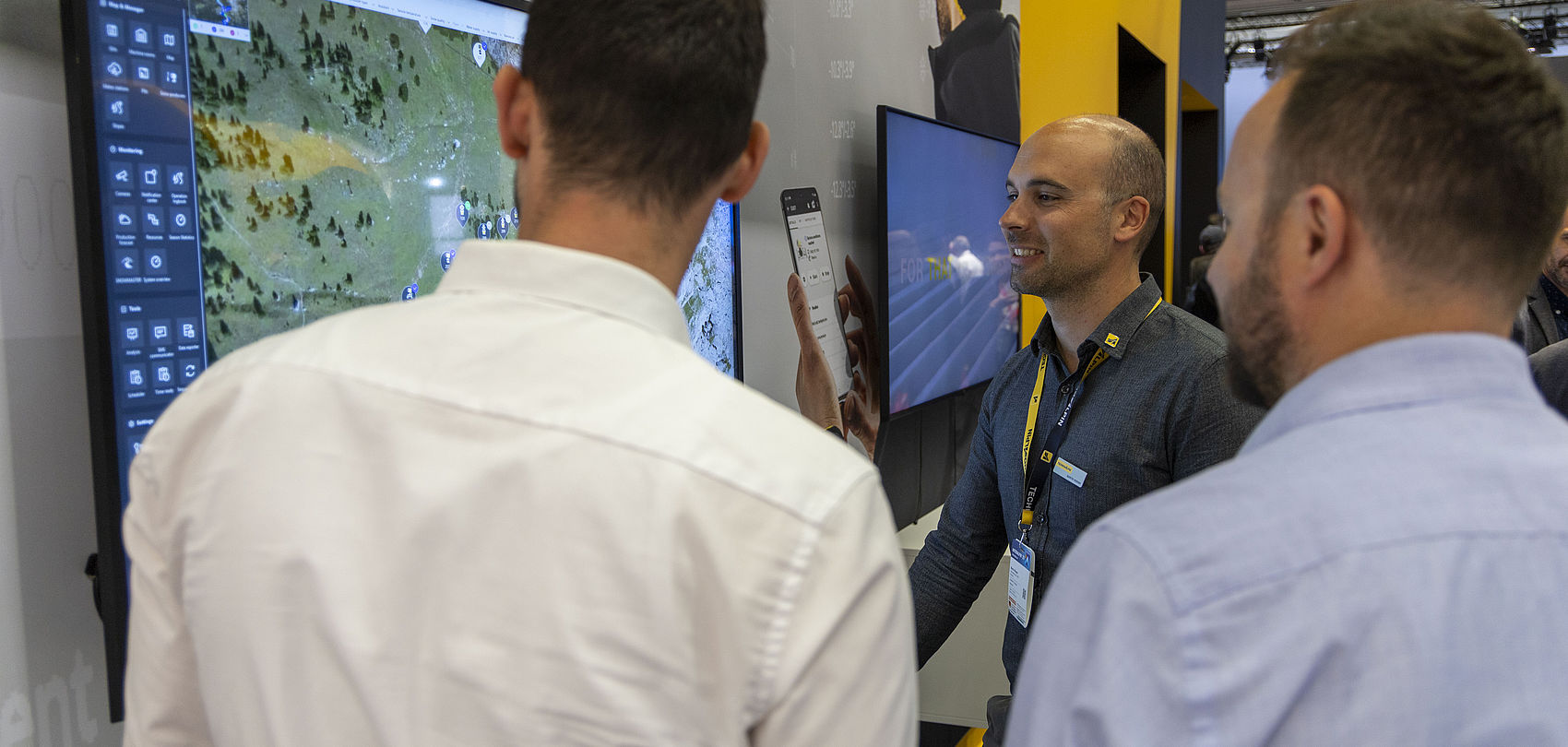 Three men at trade fair booth looking at screen with map and software application