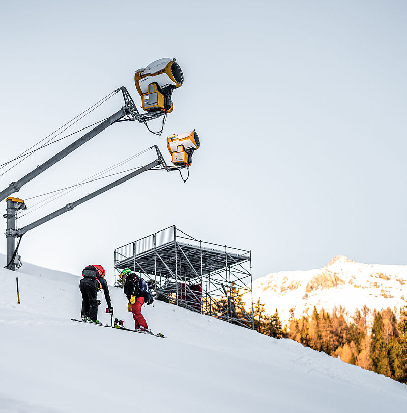 Two skiers on slope in front of TechnoAlpin snow guns and ski jump in Crans Montana