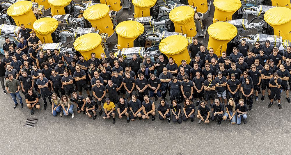 Großes Gruppenfoto der TechnoAlpin Mitarbeiter in schwarzen Shirts vor gelben Schneekanonen