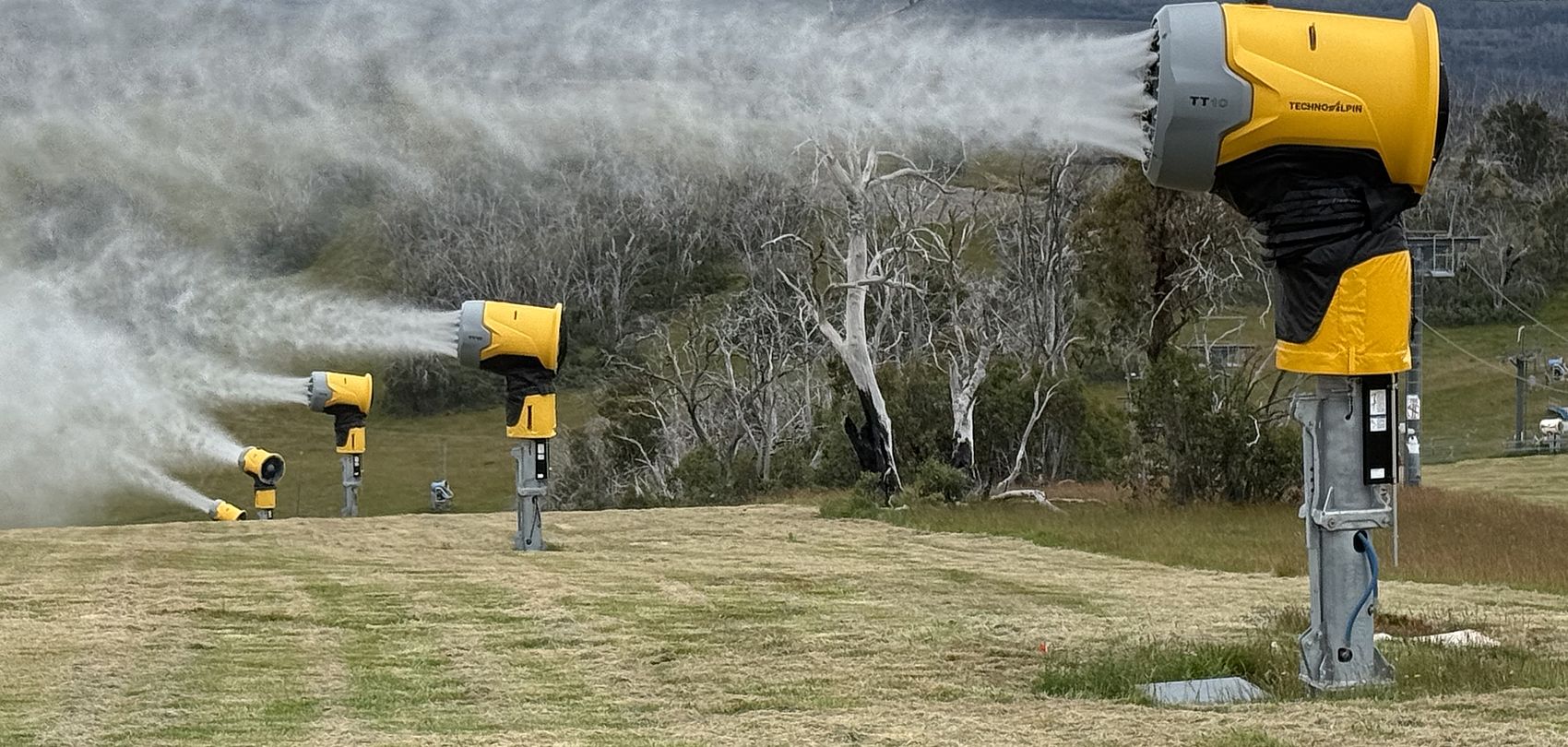 Several yellow TechnoAlpin TT10 snow guns spraying water over green summer meadow
