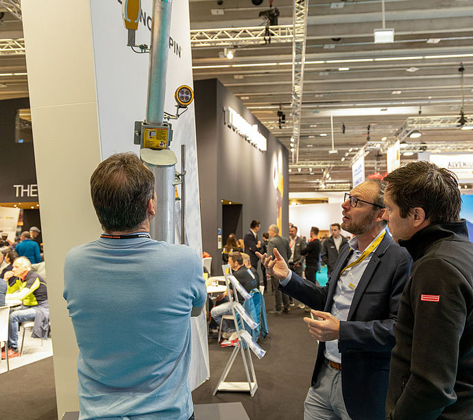 Zwei Männer in Businesskleidung diskutieren Schneelanze am TechnoAlpin Interalpin 2023 Stand, weiterer Besucher begutachtet das Gerät