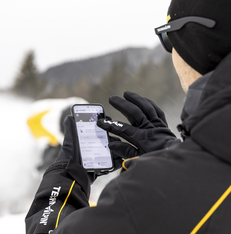 Techniker in TechnoAlpin-Jacke bedient ATASSpro Steuerung auf Smartphone in verschneiter Bergkulisse