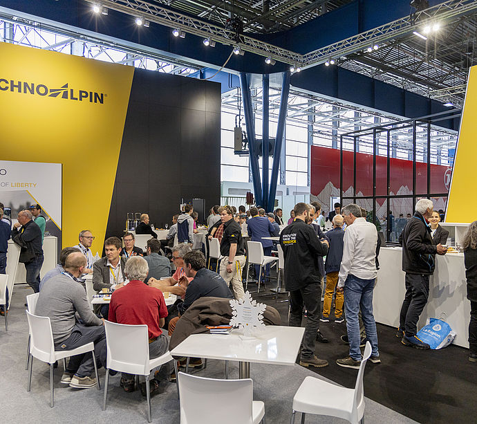 Busy TechnoAlpin trade show stand with yellow-black branding and networking area.