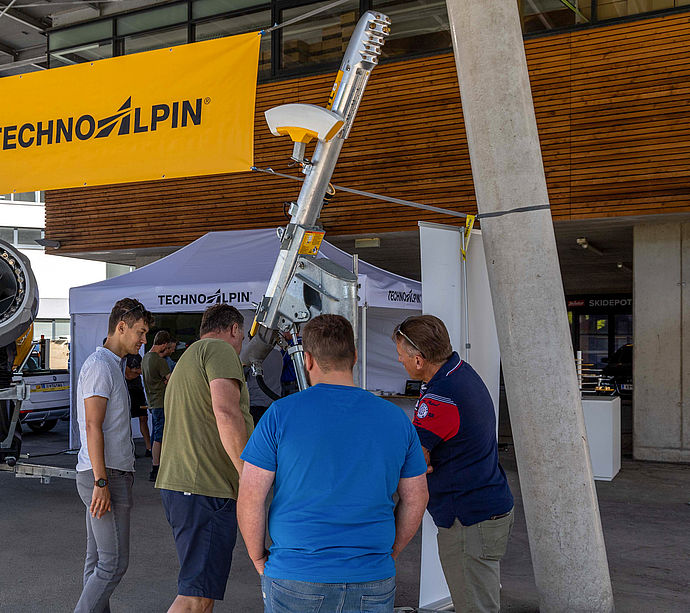 Visitors inspecting snow lance under TechnoAlpin banner in front of wooden facade.