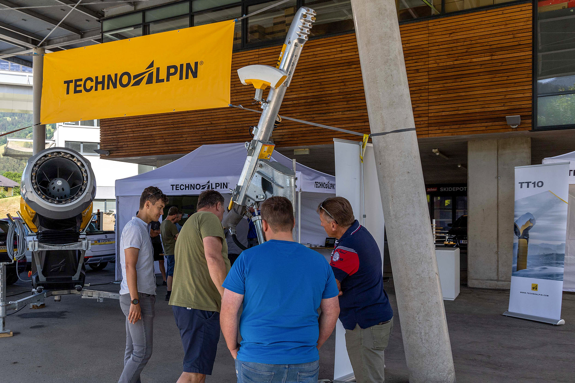 Visitors inspecting snow lance under TechnoAlpin banner in front of wooden facade.
