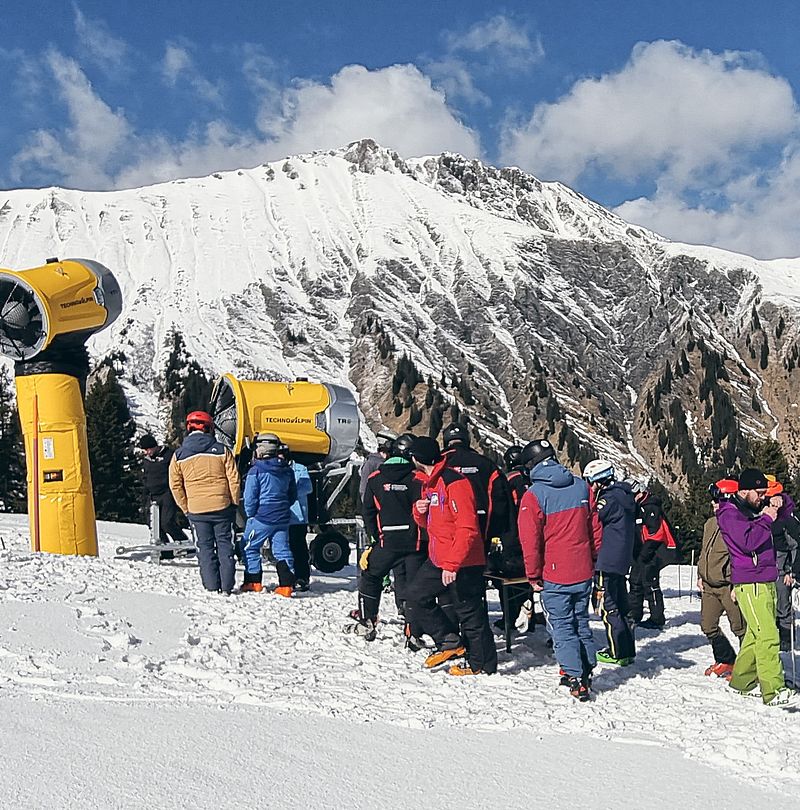 Gruppe von Personen begutachtet eine gelbe TechnoAlpin TR-Schneeerzeuger auf verschneiter Piste vor Bergpanorama
