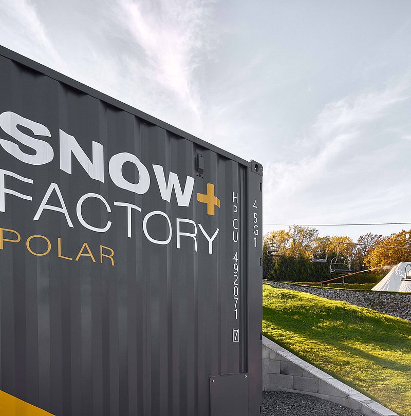 Container SnowFactory Polar grigio-giallo su prato verde con cumulo di neve e seggiovia