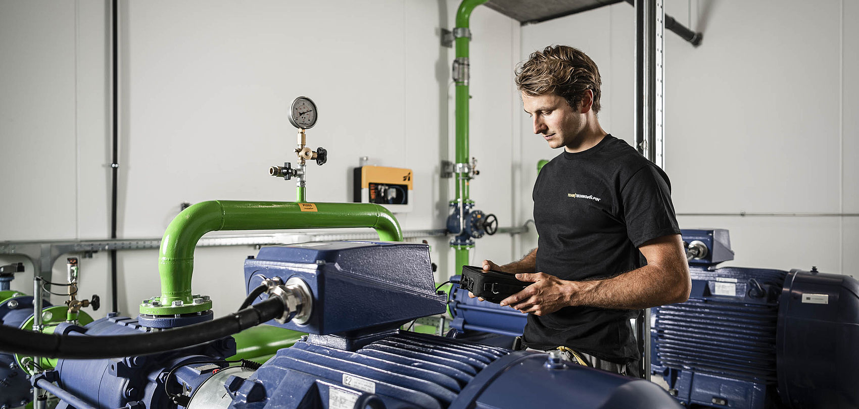 TechnoAlpin Techniker im schwarzen Shirt prüft mit Messgerät blaue Pumpen und grüne Rohrleitungen