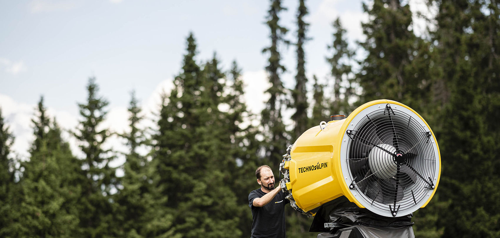 Technicien monte enneigeur à hélice TechnoAlpin jaune devant forêt de conifères
