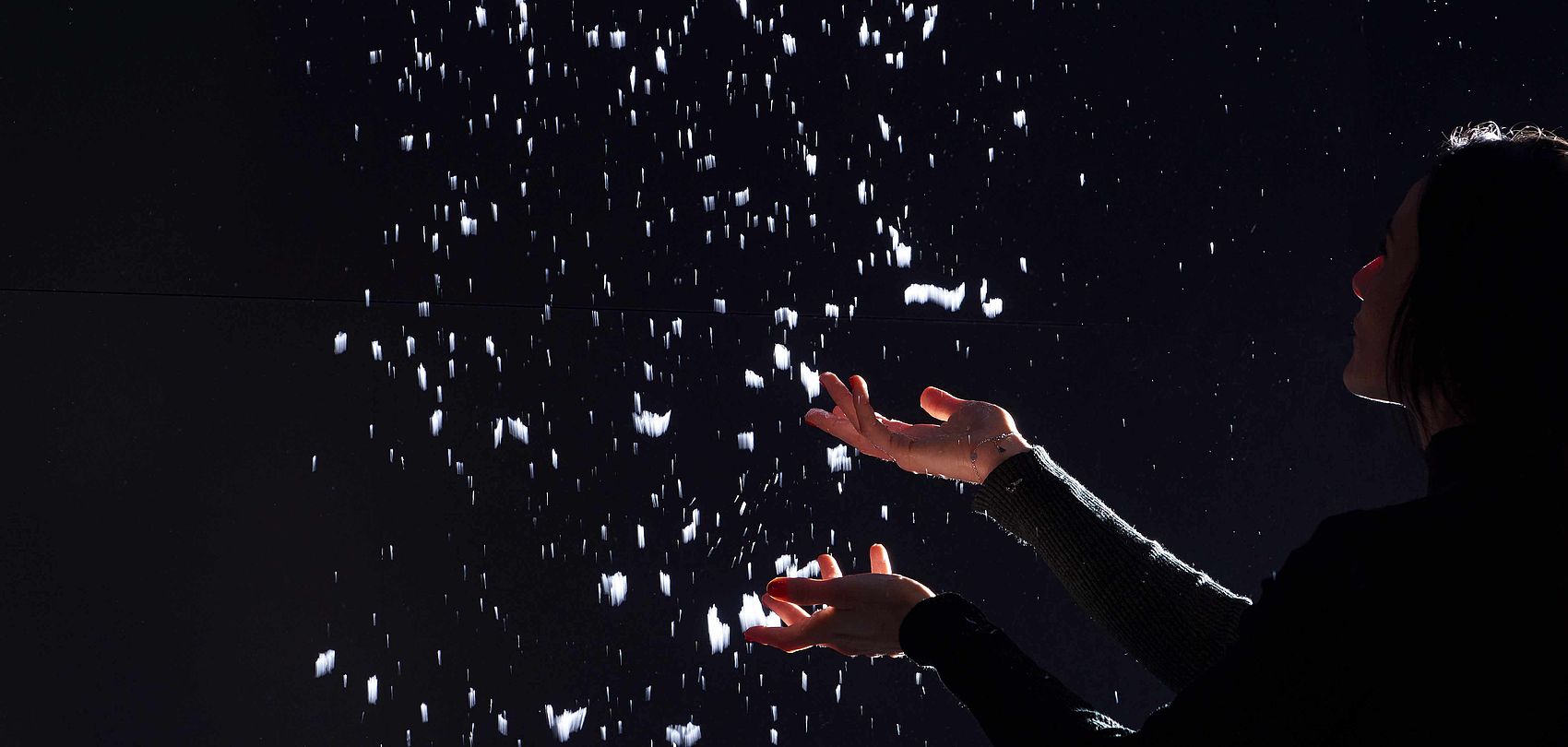 Silhouette einer Person, die mit erhobenen Händen fallende Schneeflocken vor dunklem Hintergrund auffängt