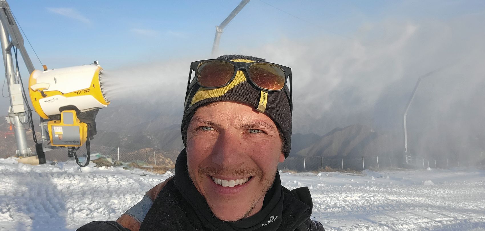 Technicien TechnoAlpin souriant prenant un selfie devant le générateur TF10 en montagne chinoise.