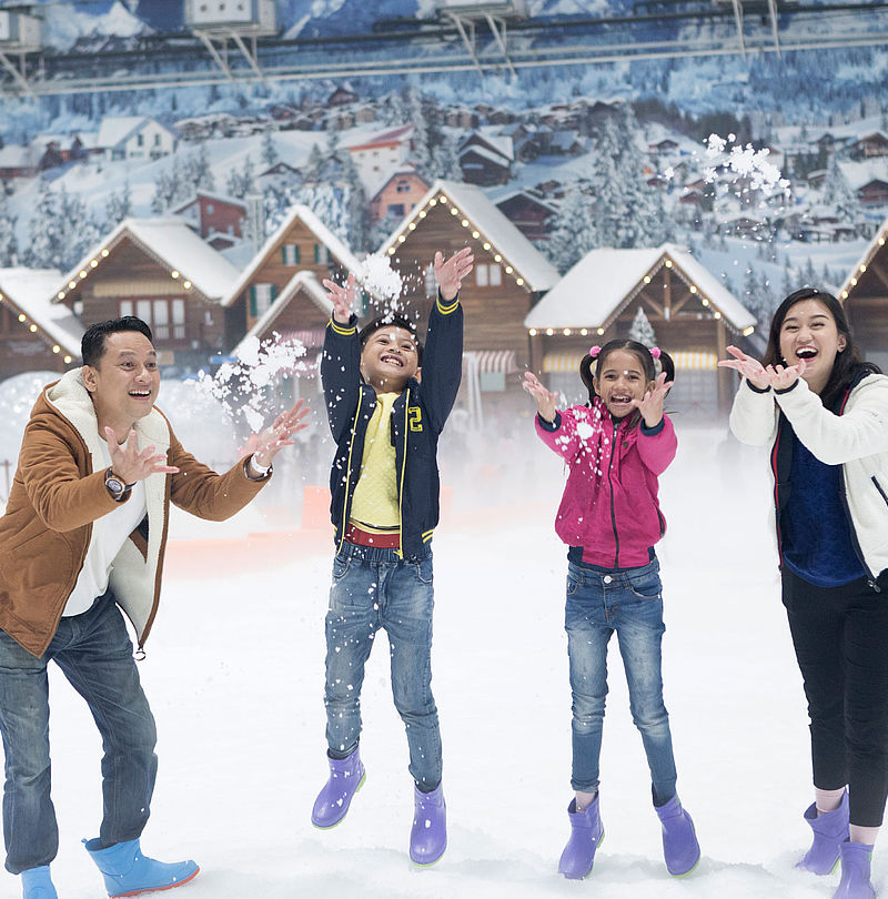 Lachende Familie wirft Schnee in die Luft vor Chalet-Kulisse in Indoor-Schneehalle