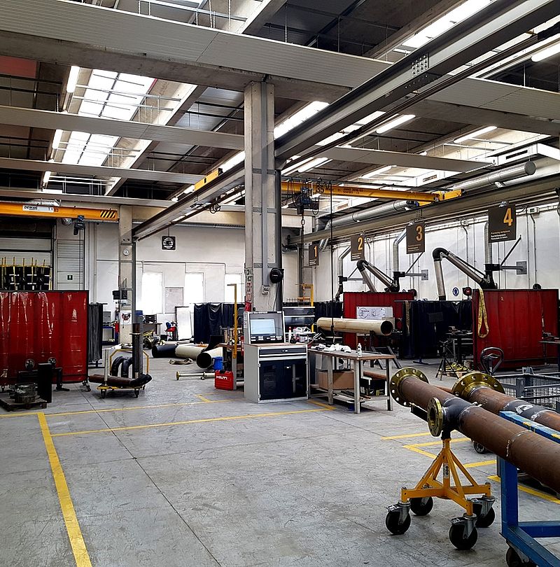 Spacious workshop with crane rails, welding stations and pipes on transport frames.
