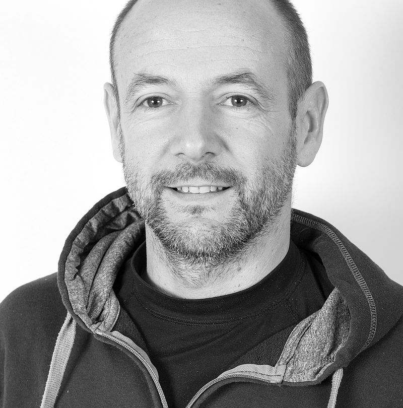 Louis Pascal Vesy, TechnoAlpin Mitarbeiter, Schwarz-Weiß-Portrait mit kurzem Haar, kurzem Bart und dezentralem Lächeln in dunklem Kapuzenpullover