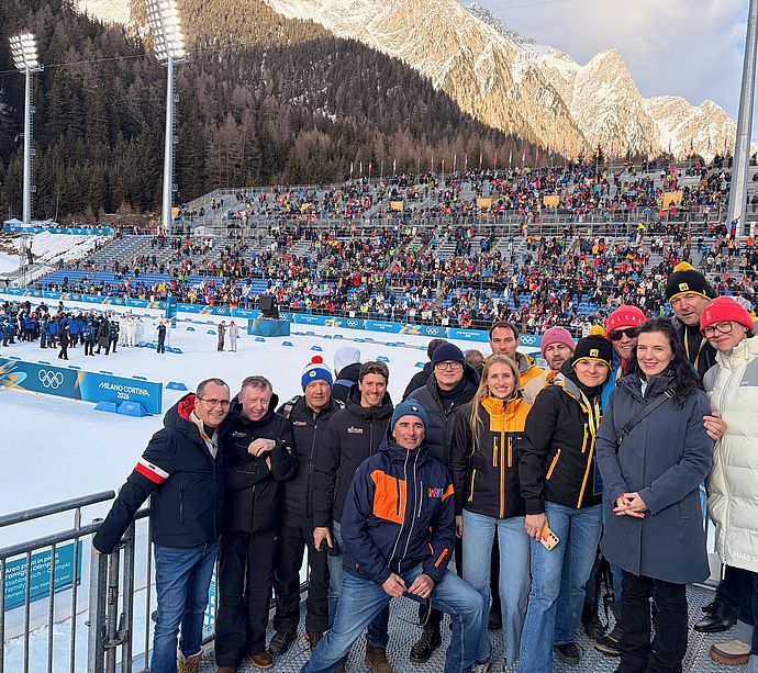 Group photo in Anterselva biathlon stadium with grandstands and snowy Dolomites at Olympics 2026
