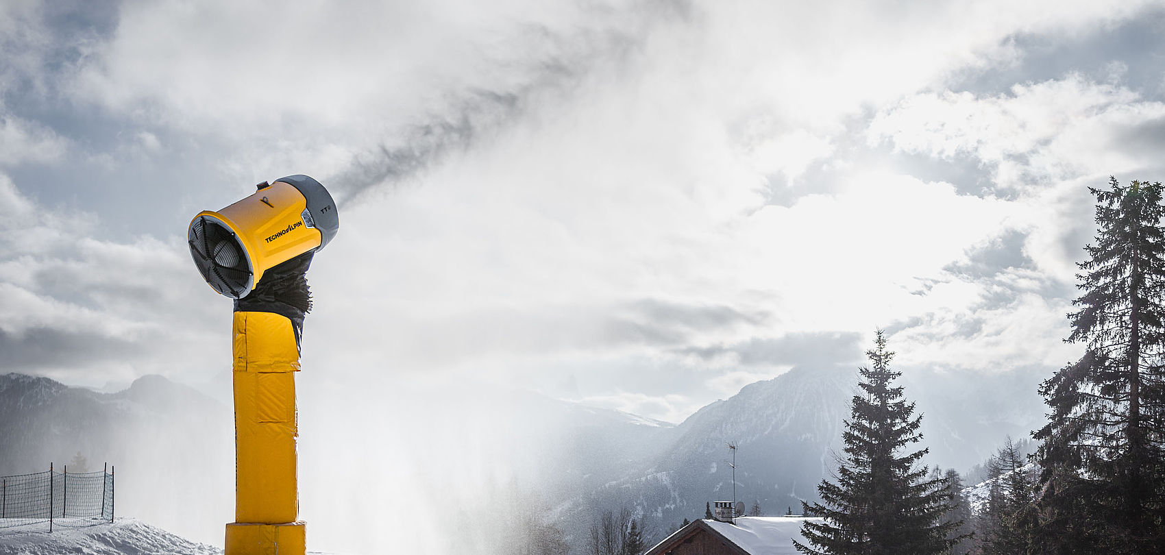 Générateur de neige TechnoAlpin TT9 jaune sur tour en activité devant paysage montagneux nuageux.
