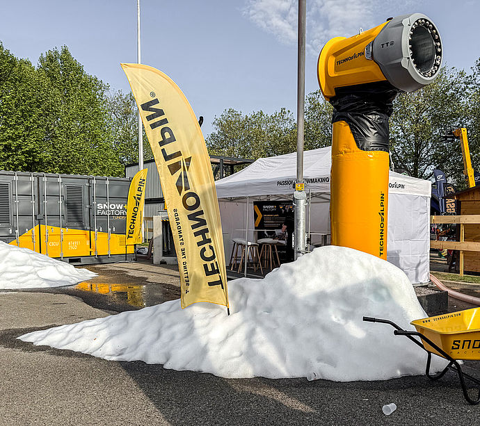 Gelber TT Schneeerzeuger im Außenbereich mit Schneehaufen, TechnoAlpin Fahnen und SnowFactory Container