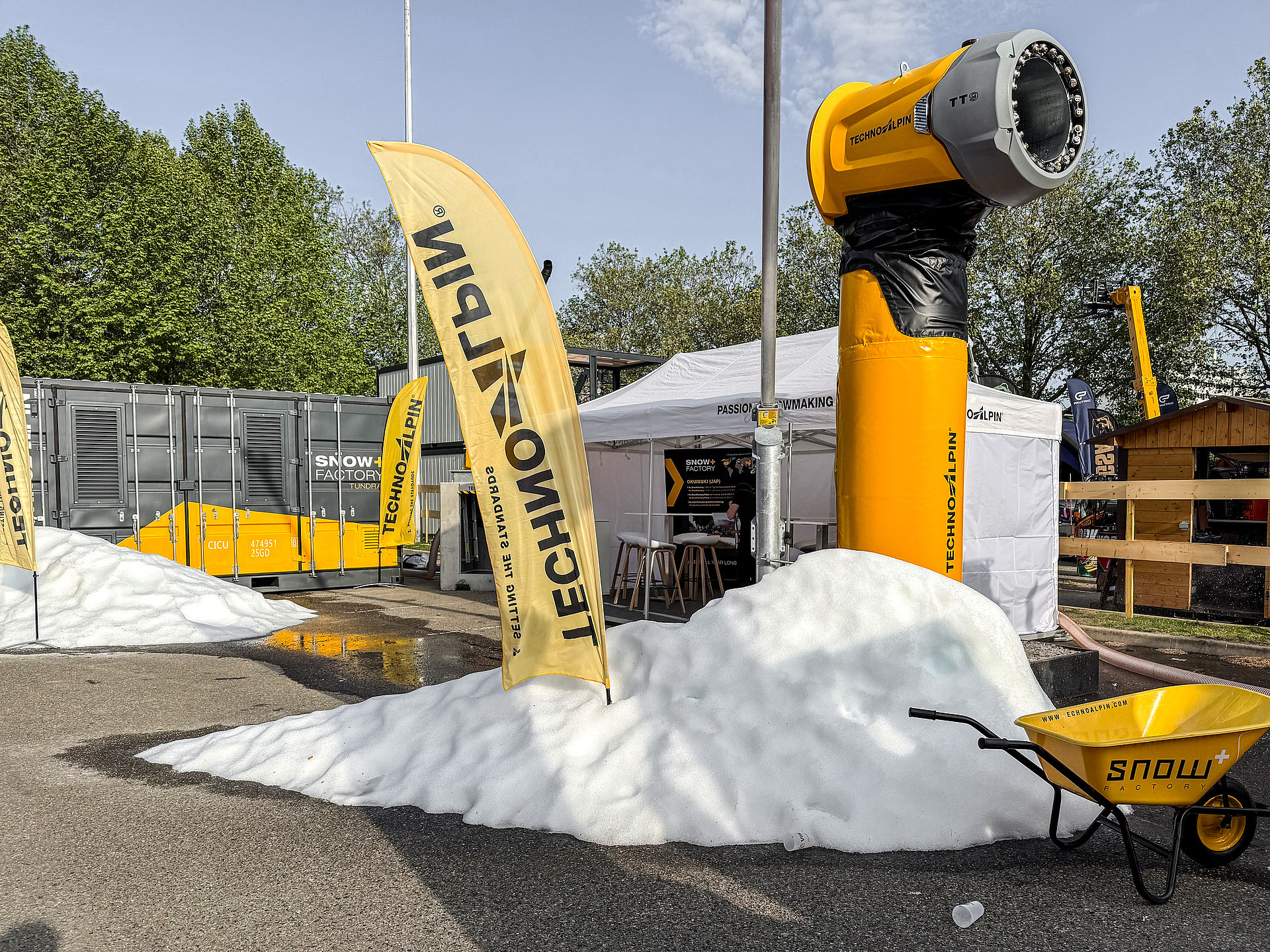 Gelber TT Schneeerzeuger im Außenbereich mit Schneehaufen, TechnoAlpin Fahnen und SnowFactory Container