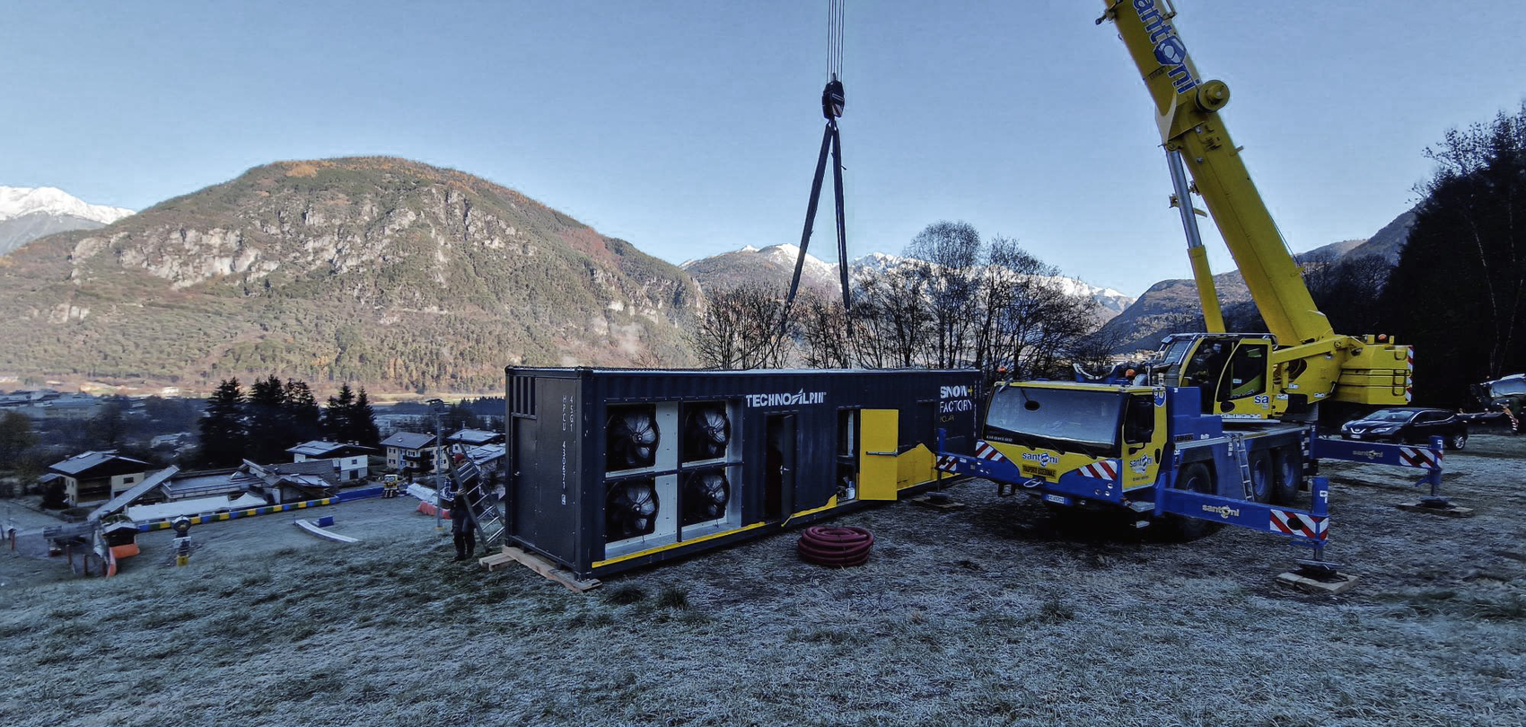 Mobilkran hebt einen TechnoAlpin Snowfactory-Container auf einer Wiese mit Bergen im Hintergrund