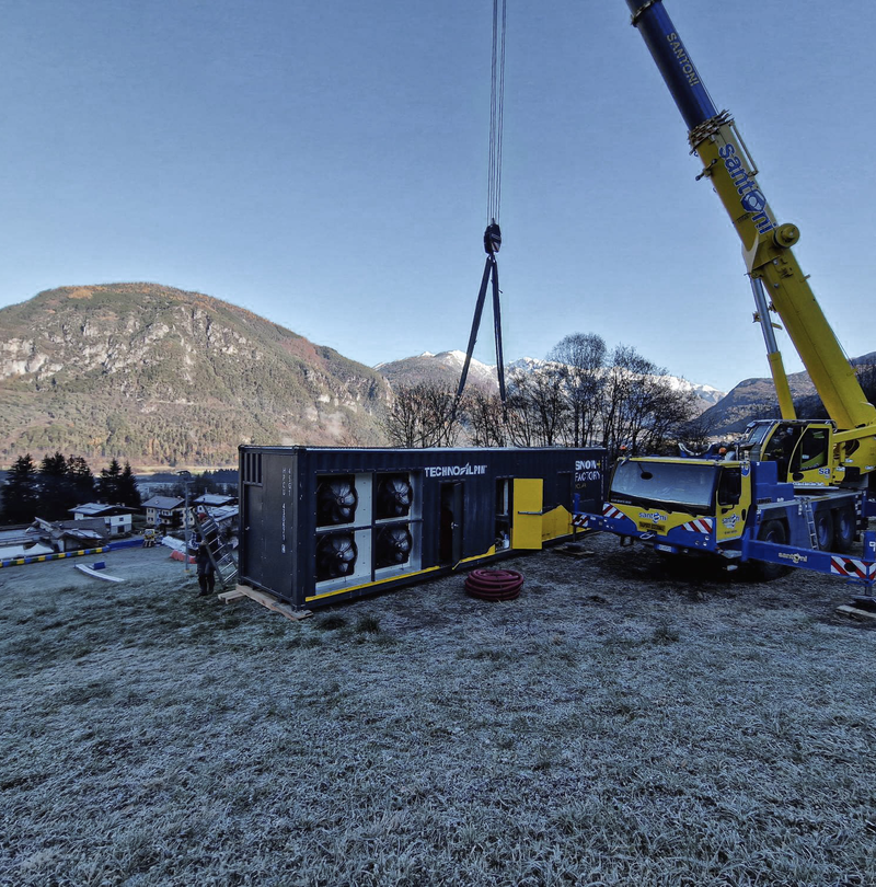 Mobilkran hebt einen TechnoAlpin Snowfactory-Container auf einer Wiese mit Bergen im Hintergrund