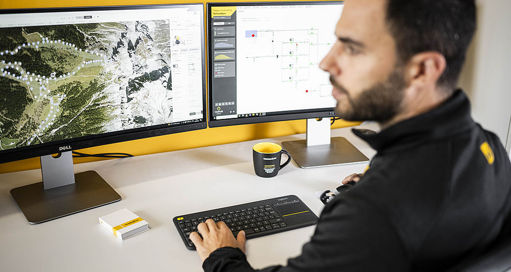 TechnoAlpin Techniker arbeitet am Doppelmonitor mit Skigebiets-Karte und Steuerungssoftware