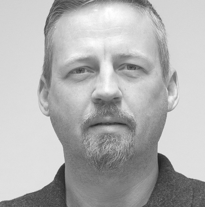 Günther Muglach, TechnoAlpin Mitarbeiter, Schwarz-Weiß-Portrait mit kurzem Haar, Ziegenbart und ernstem Blick im dunklen Hemd