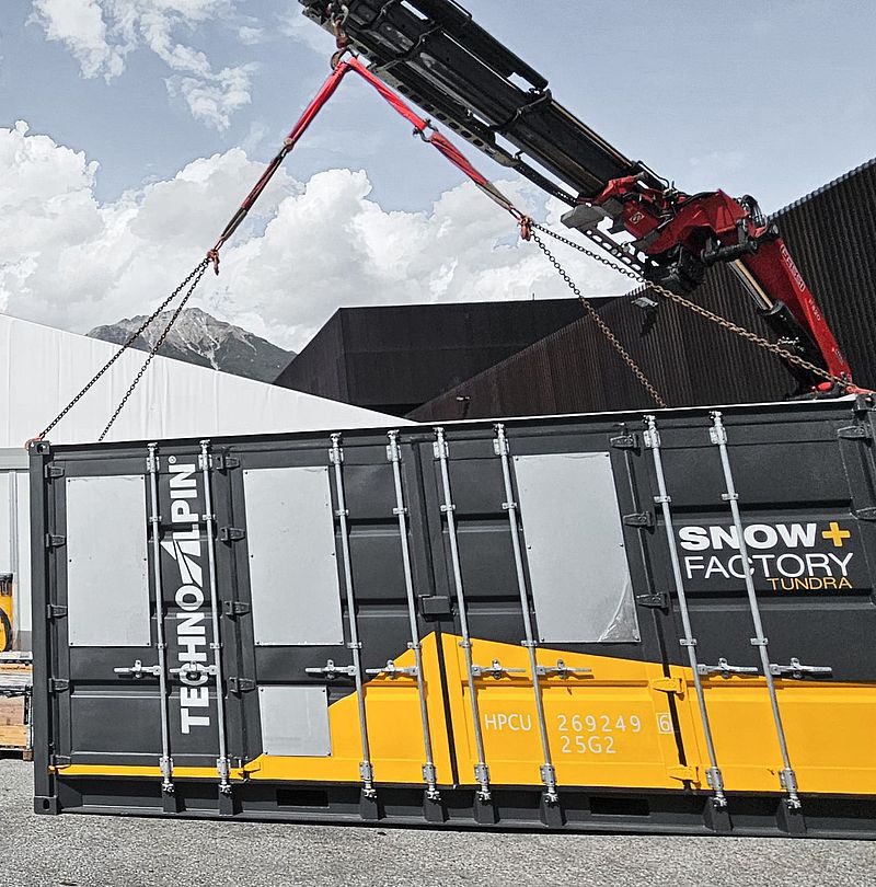 Crane loads TechnoAlpin Tundra SnowFactory container in front of mountain panorama