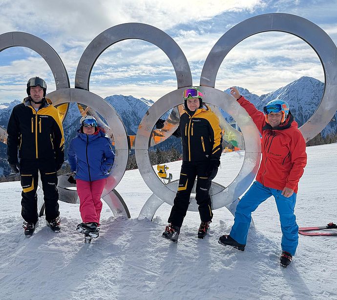 Four people in ski clothing pose in front of Olympic rings on snowy mountain peak