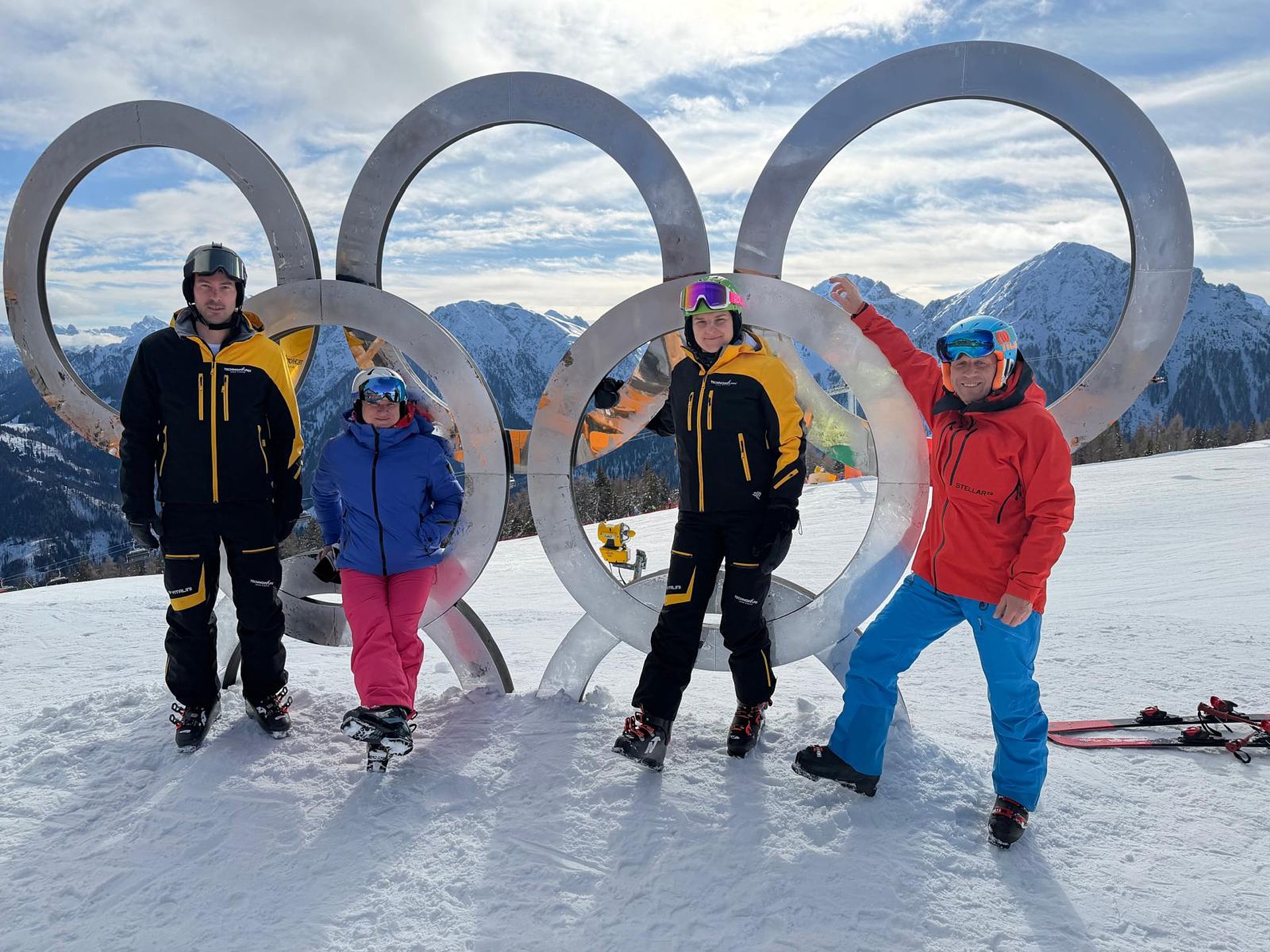 Four people in ski clothing pose in front of Olympic rings on snowy mountain peak