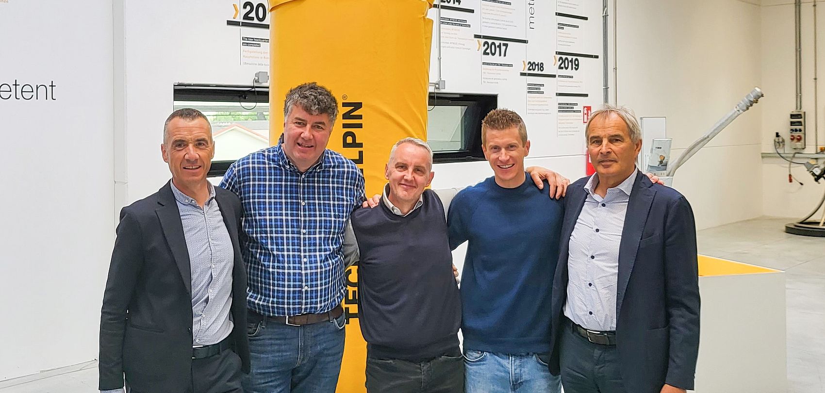 Five men in group photo in front of yellow TechnoAlpin pillar and company timeline.