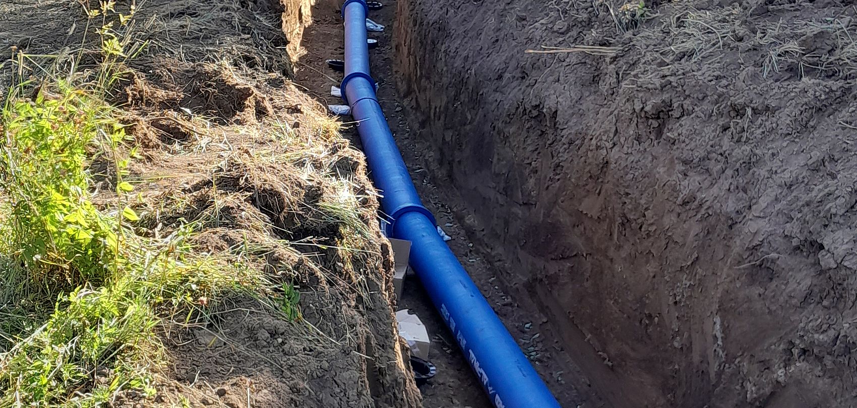 Blue Alpinal snowmaking pipes laid in a long excavated trench running through a grassy hillside lined with trees, pipeline infrastructure