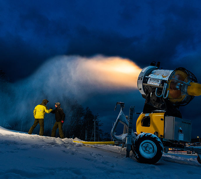 Two people next to mobile propeller snow cannon at dusk