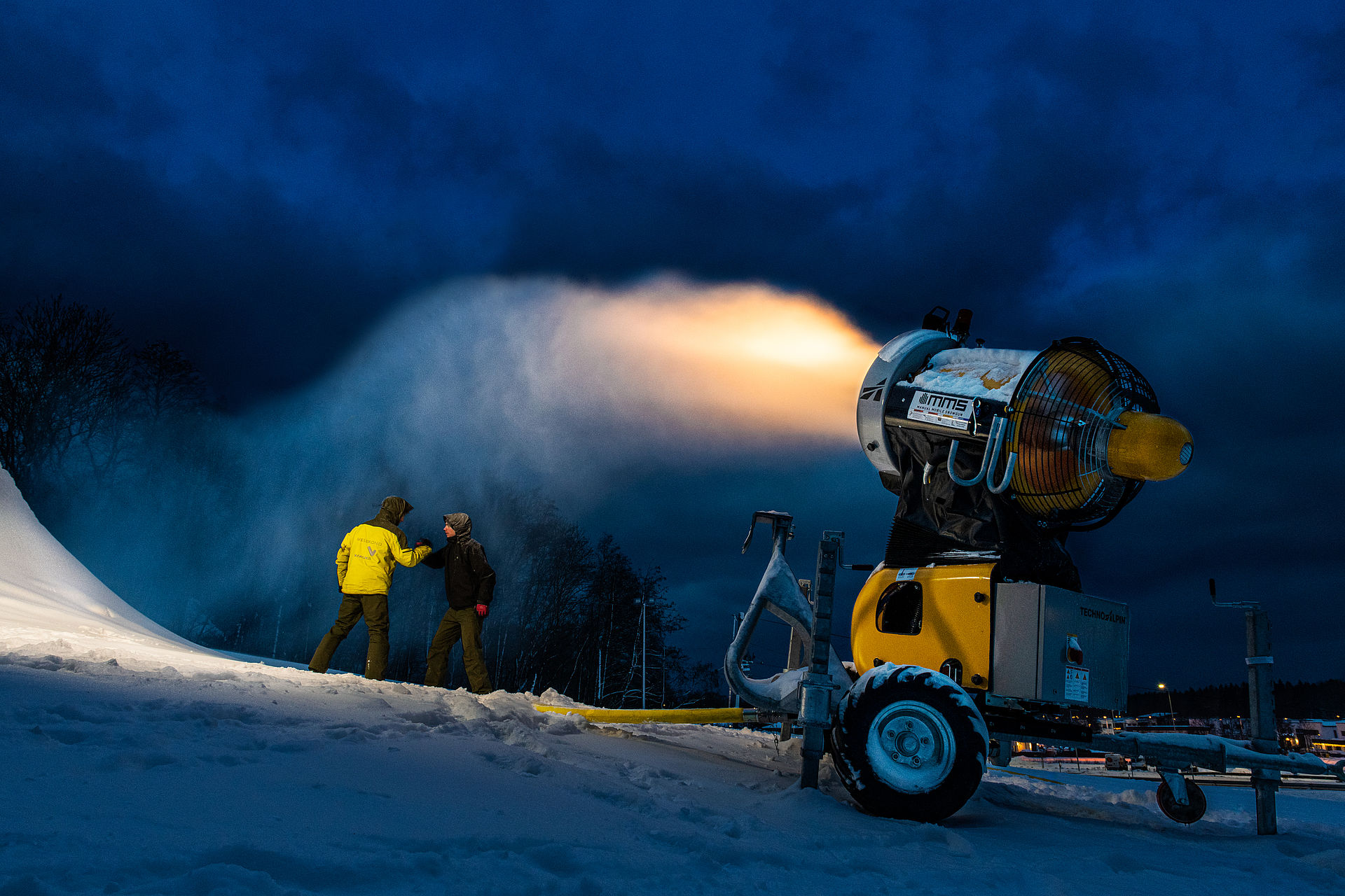 Two people next to mobile propeller snow cannon at dusk