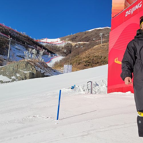 Manuel Schöpf in schwarzer Jacke und Mütze lächelt am Zielbereich einer Skirennen-Strecke bei den Olympischen Spielen in Peking