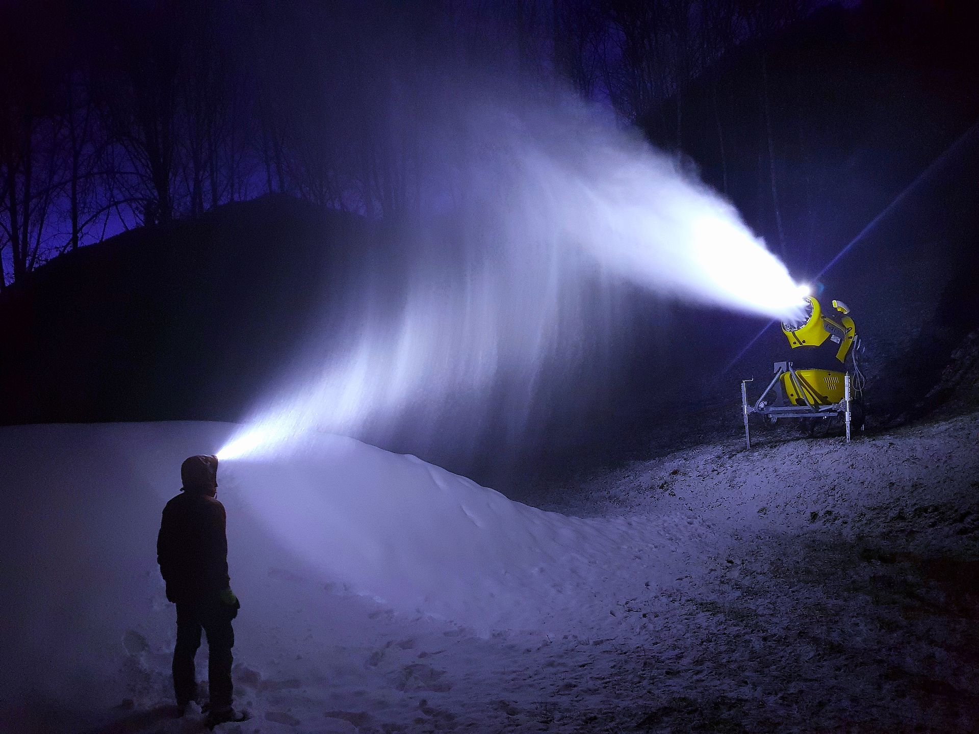 Persona con lampada frontale osserva di notte un cannone da neve TechnoAlpin durante l'innevamento