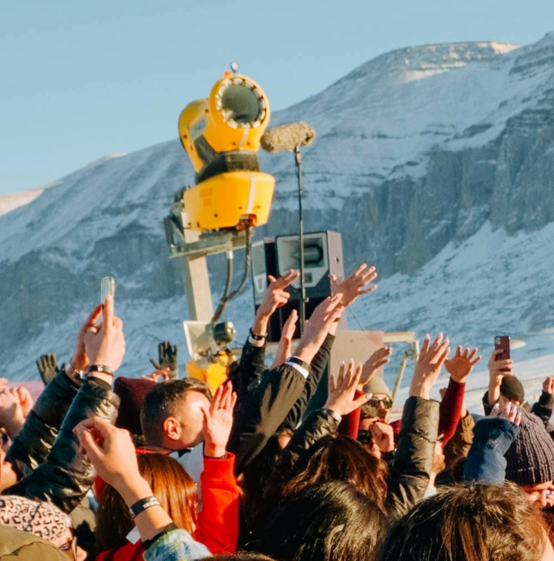 Foule avec mains levées lors d'un événement devant enneigeur TechnoAlpin et paysage montagneux enneigé