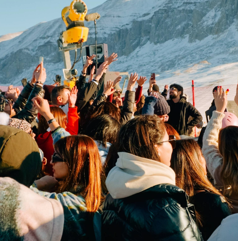 Crowd with raised hands at event in front of TechnoAlpin fan gun and snowy mountain scenery