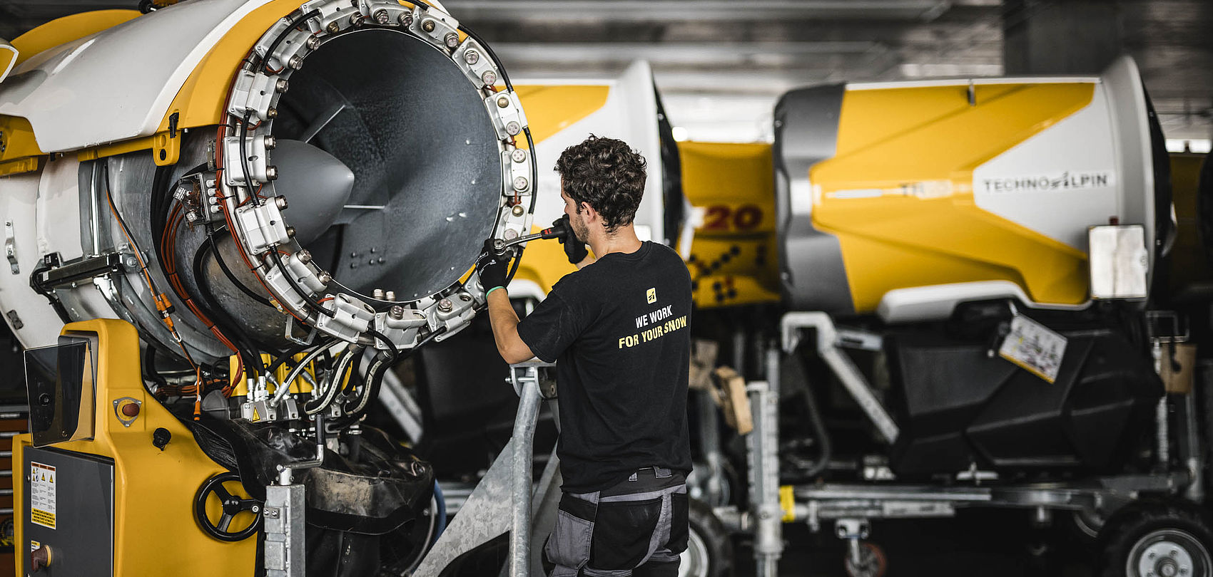 Technician in black T-shirt works on yellow-white TechnoAlpin snow gun in workshop
