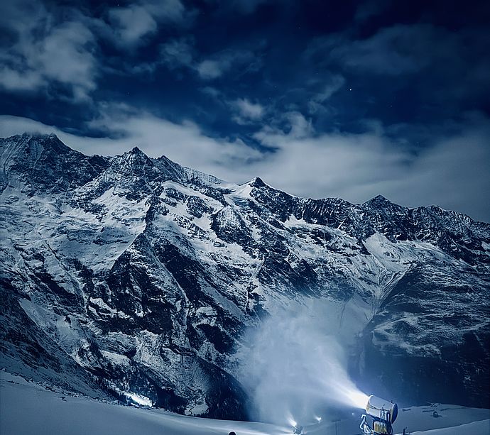 Person mit Schaufel vor Schneekanone bei Nacht vor schneebedeckter Berglandschaft