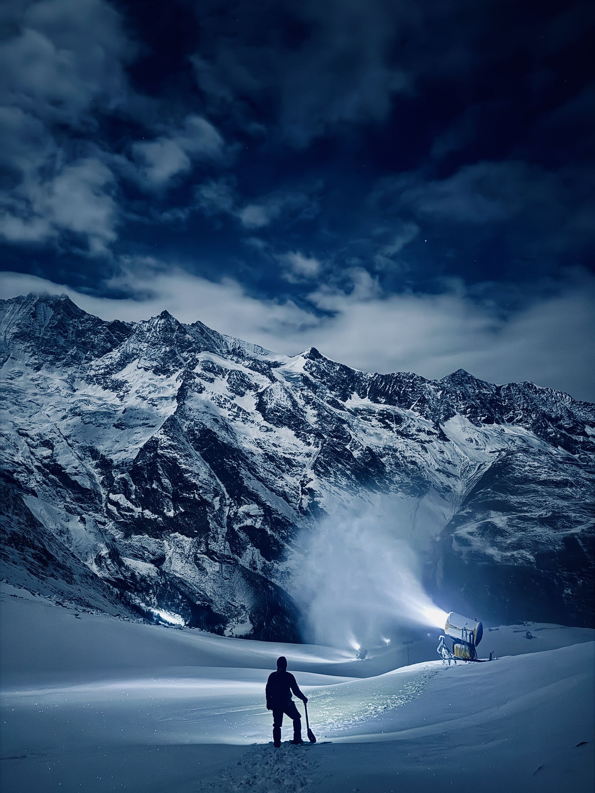 Person mit Schaufel vor Schneekanone bei Nacht vor schneebedeckter Berglandschaft