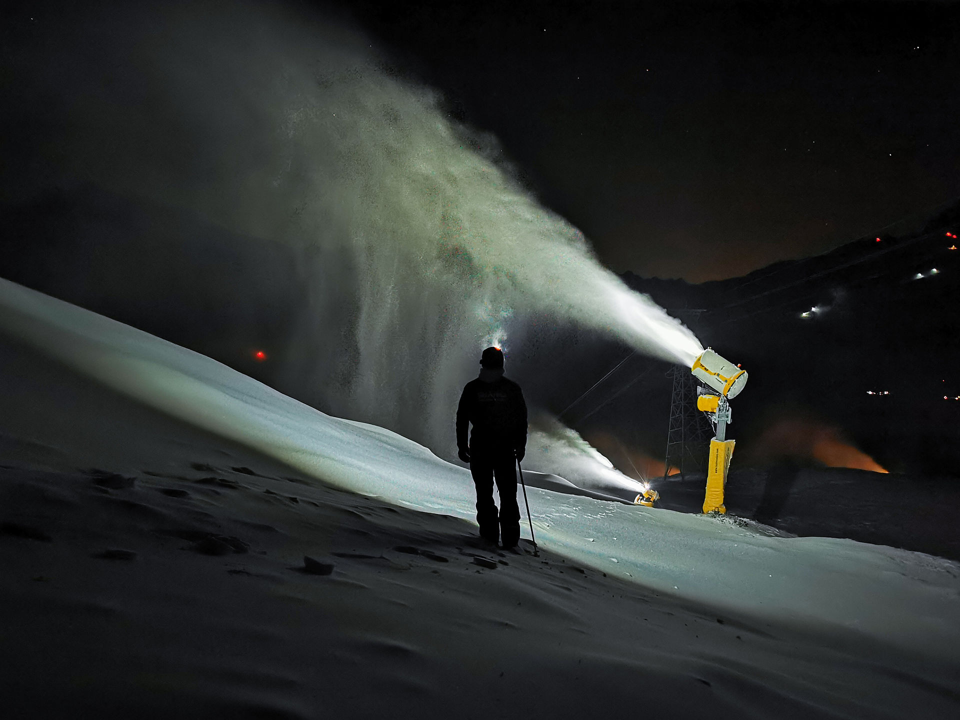 Nighttime snowmaking with person in front of illuminated snow cannon
