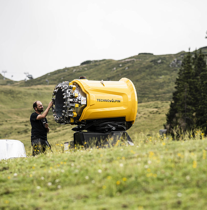Tecnico TechnoAlpin ispeziona corona ugelli aperta di cannone da neve giallo su prato alpino verde