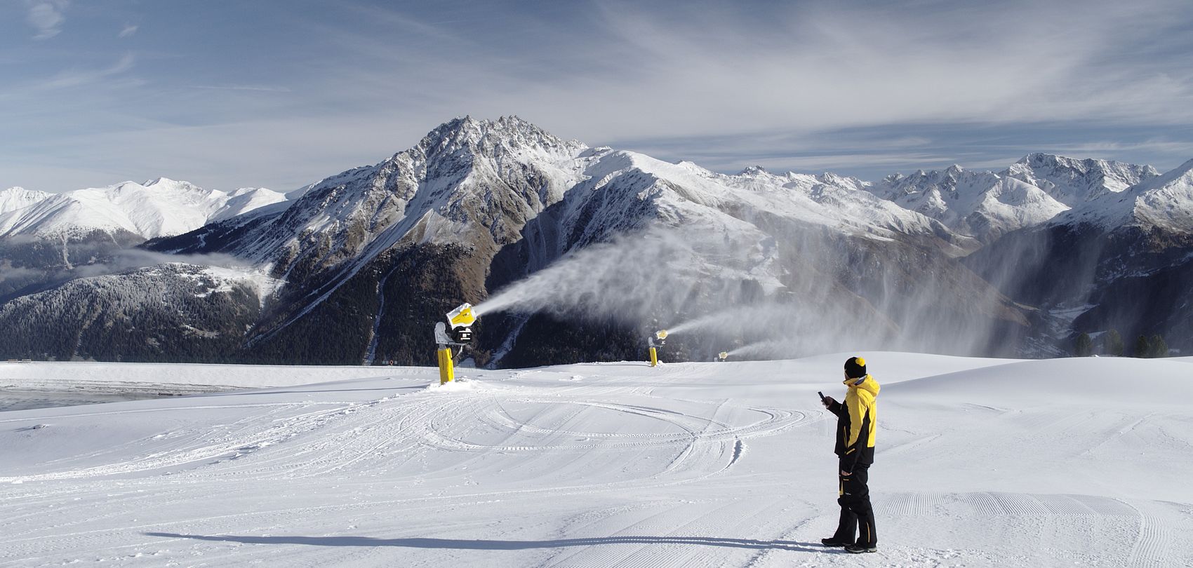 Employee with smartphone controlling snow generator on groomed slope in front of alpine panorama.