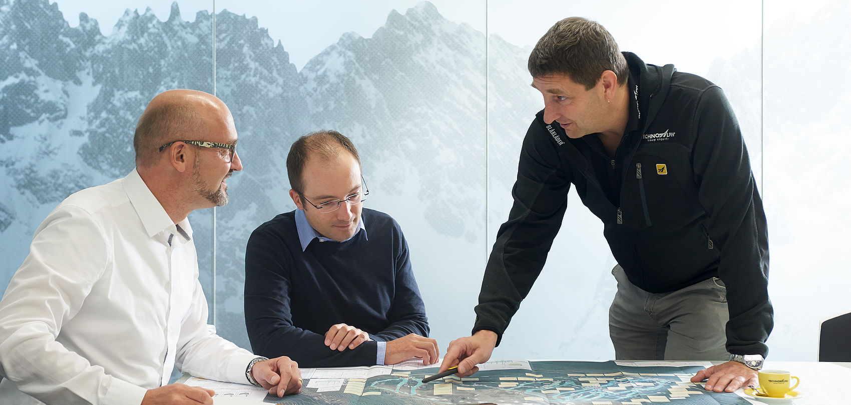 Trois hommes en consultation à table avec plans et tablette devant mur avec motif montagneux.