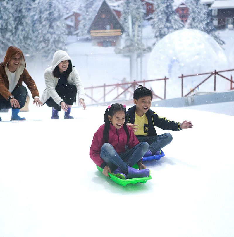 Lachende Kinder beim Schlittenfahren auf Indoor-Schneepiste mit Eltern im Hintergrund