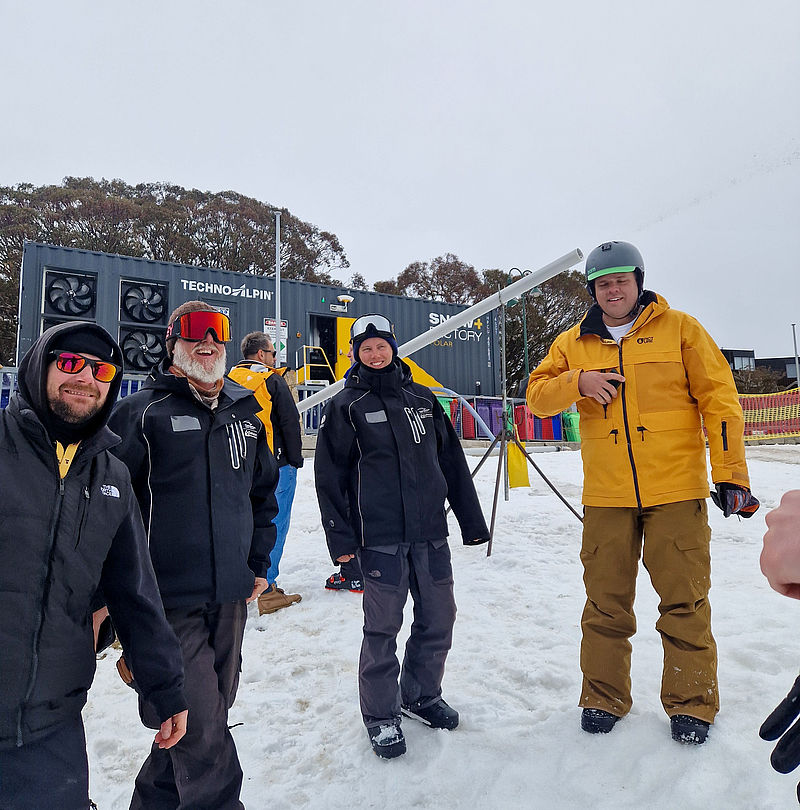 Four smiling people in winter clothing in front of TechnoAlpin SnowFactory container in snow